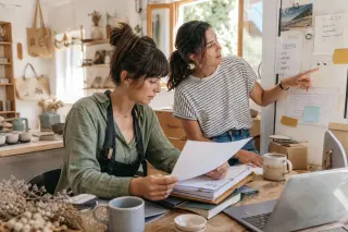 Two entrepreneurs in a naturally lit artisan workshop collaborating over documents and tax notes, with a warm and authentic atmosphere.