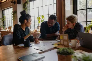 Three entrepreneurs collaborating at a wooden table in a warm coworking café, focused on notes and prototypes, sunlight creating a natural and inviting setting.