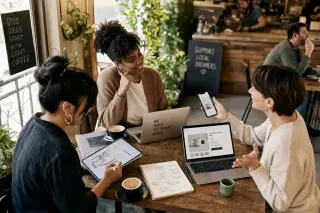 Three entrepreneurs in a sunlit café, collaborating with digital devices around a wooden table, engaged in a natural, focused exchange