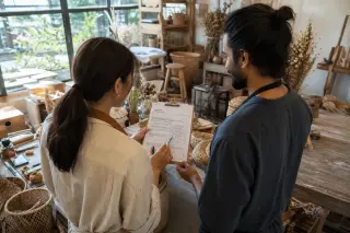 Two artisans in a bright workshop reviewing documents amid handmade products and tools, natural light highlighting their authentic collaboration.