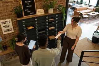 Three entrepreneurs in a modern, plant-filled coworking space collaborate around shared mailboxes, engaging with both physical letters and a digital tablet interface under warm natural light.