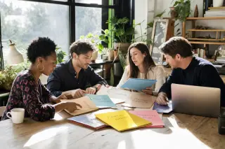 Several entrepreneurs collaborate in a warm, daylight-filled café workspace, exchanging advice with documents and folders on a shared table, immersed in a candid, natural discussion.