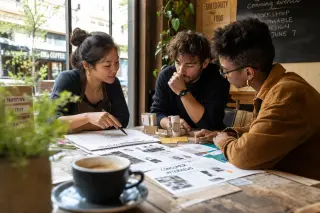 Three entrepreneurs collaborating around a café table with laptops, sketches, and notes, in warm natural light, suggesting teamwork on funding strategies.